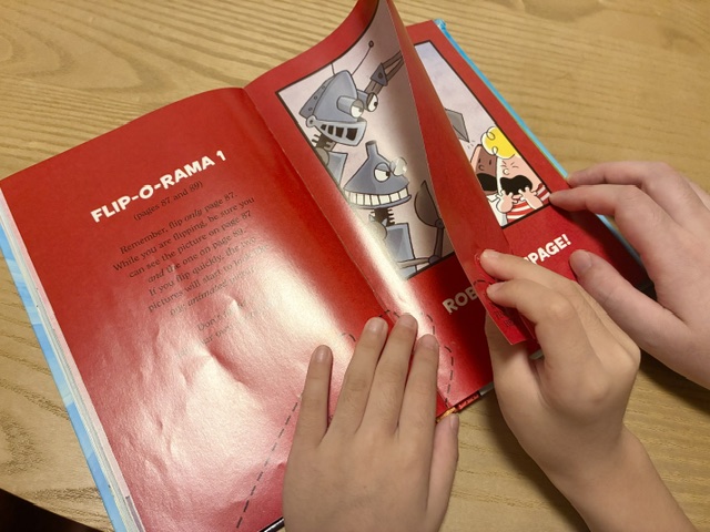 Captain UnderpantsのFlip-O-Ramaのページを指ではさんで父と一緒にめくる息子の手元。